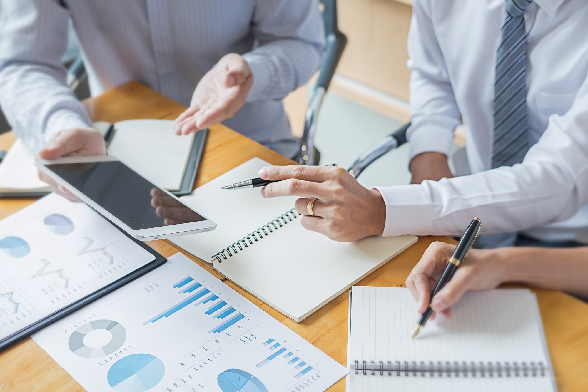 Hands of business professionals discussing data on a tablet and printed charts during a meeting. Clear focus on collaboration and analysis.