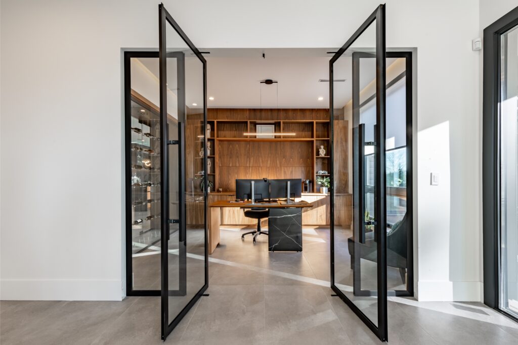 Modern home office with large open glass doors, a sleek wooden desk, dual monitors, and a black chair. Bright, minimalist design with a warm tone.