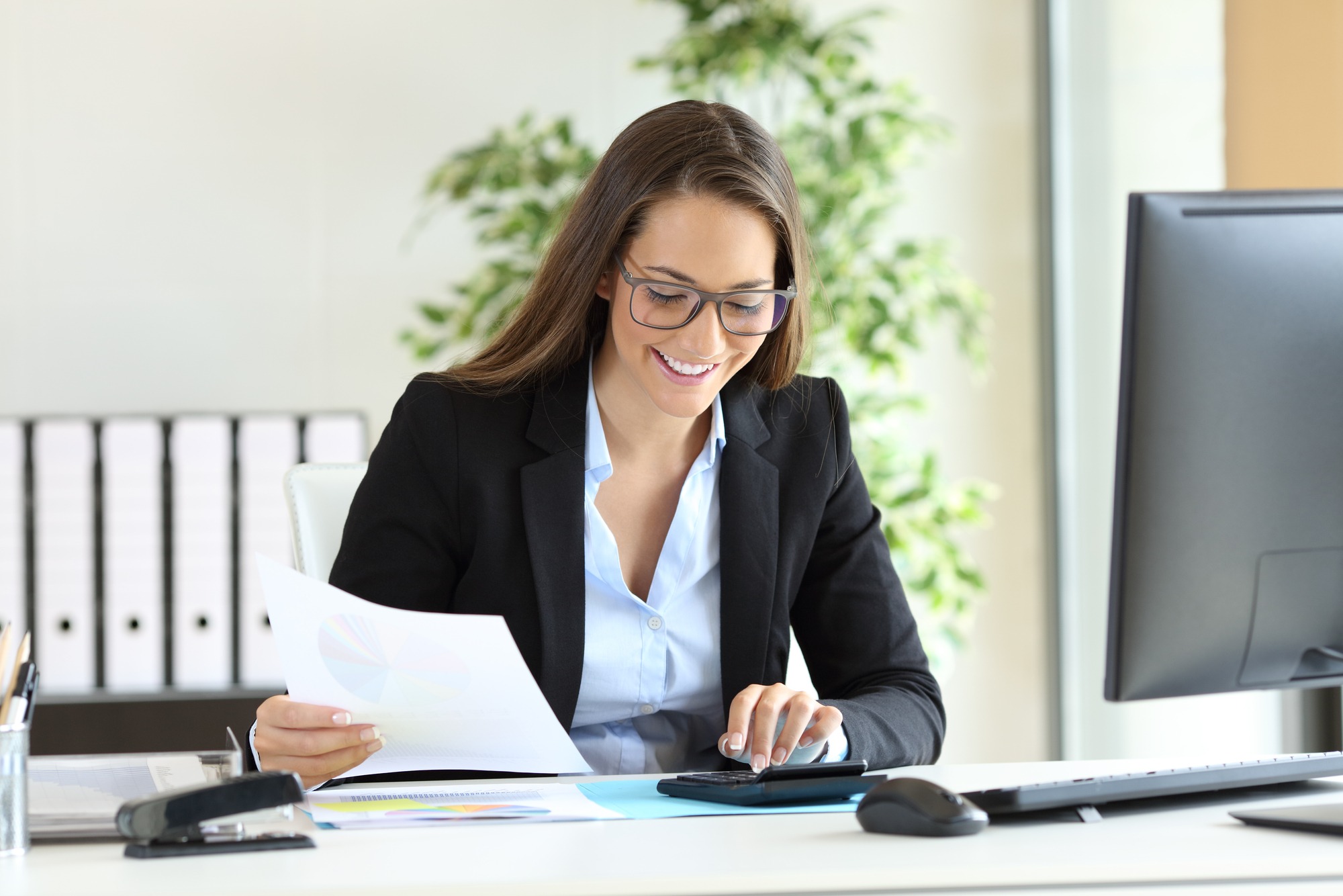 A professional woman in a black suit reviews documents and uses a calculator at her desk, focusing on her work in a bright office space.