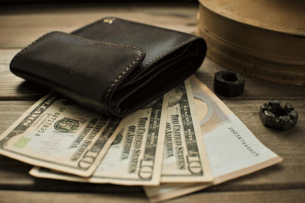 A brown leather wallet rests on a stack of cash, including several $10 bills, with metal nuts nearby against a wooden surface.